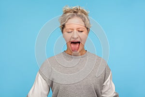 Naughty woman with short hair in sweatshirt standing with closed eyes and sticking out tongue