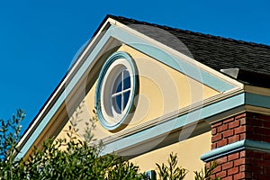 Nautical round window in attic room at pinnacle of home with beige stucco exterior and gable style roof white accents