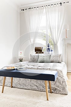 Navy blue bench with book and breakfast standing by the king-size bed in white bedroom interior with window with curtains