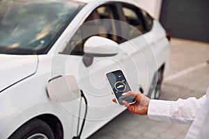 Near garage. Close up view. Woman charging her electric car and monitoring process on smartphone