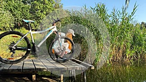 Near the lake on a bicycle, a girl looks into water while standing on a bridge.