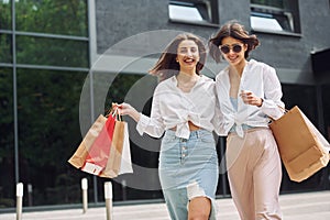 Near modern building. Two female friends have a shopping day. Walking outdoors with bags