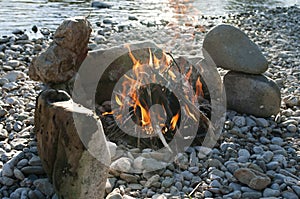 Near the river campfire ablaze with stone circle. Camping season, summer, joy