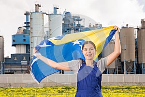 near suburban factory,cheerful female worker in overalls stands and holds Bosnian flag in her hands