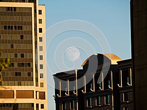 Nearly Full Moon In Blue Sky Above Buildings