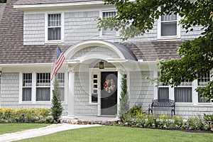 Neat and pretty shingled retro house with arched entryway and beautiful landscaping with colorful summer wreath on front door and