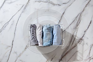 Neatly folded clothes and pyjamas in the metal mesh organizer basket on white marble table.