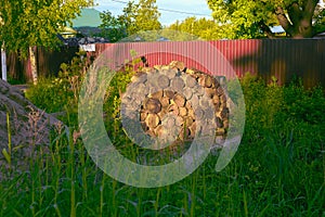 The neatly woodpile made of sawn tree trunks among the grass in the rays of the rising sun, near a metal tinted fence