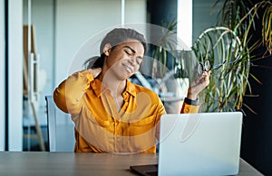 Neck ache. Black businesswoman massaging aching neck, suffering from pain after computer work, sitting in office