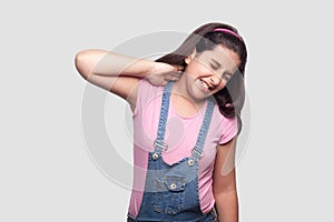 Neck pain. Portrait of sad brunette young girl in pink t-shirt and blue overalls standing and holding her painful neck and feeling