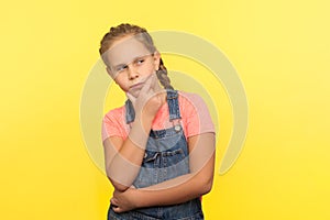 Need to think! Portrait of clever pensive little girl in denim overalls holding chin and pondering solution