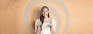 We need you. Smiling young woman pointing fingers at camera to beckon or invite people, standing on beige background