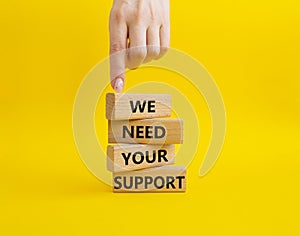 We need your support symbol. Wooden blocks with words We need your support. Beautiful yellow background. Businessman hand.