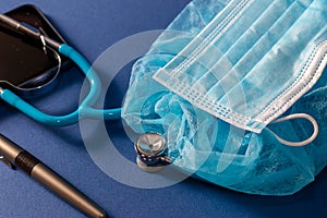 Neonatal blue stethoscope, medical cap and mask on blue dark background, pen and partial view of a cellular phone.