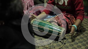 A Nepali Indian young girl in bright clothes plays a national drum of madal sitting on the floor of the room. Close-up