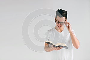 Nerd with glasses and a white t-shirt is reading a book on a white background