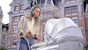 Nervous busy mom swinging infant in pram and chewing bun, having snack in park