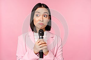 Nervous female entrepreneur giving speech with microphone, holding mic and looking aside anxiously, posing in suit over