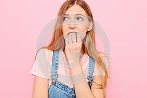 Nervous insecure scared young woman looks away, feeling afraid insecure agitated scared biting her nails, pink background