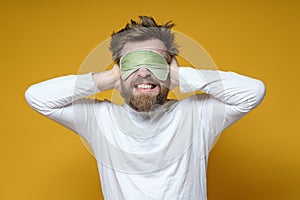 Nervous man in a sleeping mask covered ears with hands so as not to hear the noise, he is annoyed and grins teeth