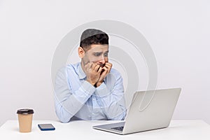Nervous scared man employee sitting office workplace, looking worried anxious at laptop screen and biting nails