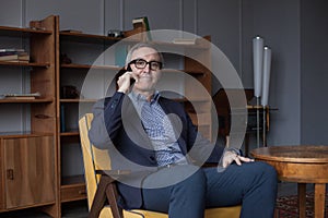 Nervous worried elderly businessman in blue suit and eye glasses talking on his mobile phone on grey wall  background