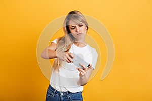 Nervous young blonde woman posing isolated over yellow wall background dressed in white casual t-shirt using mobile phone play