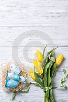 Nest with easter eggs and yellow tulips bouquet on wooden background, top view