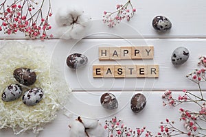 Nest with naturally dyed eggs, cotton flowers decor and and inscription Happy Easter made from wooden letters on wight wooden