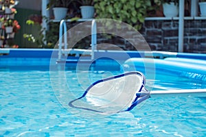 A net for cleaning the pool in the clear blue pool water. In the background is a courtyard with flowers. Round frame