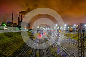 A network of intertwined railroad tracks illuminated by bright illumination at night and pipes of industrial enterprises that spew