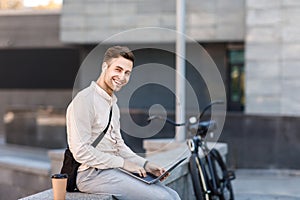 Networking outdoors. Happy guy in wireless headphones with coffee, works on laptop in city