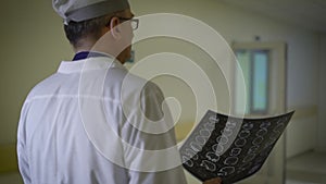 A neurosurgeon physician who is dressed in a white robe walks along the corridor of the clinic and holds the results of