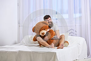 Never grow up concept. Guy on happy face hugs giant teddy bear. Man sits on bed and hugs big toy, white curtains on