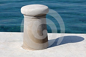 New dark grey concrete mooring bollard on top of stone and concrete pier with calm clear blue sea in background