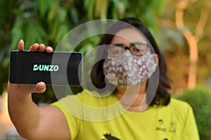 New Delhi, India, 2020. Girl wearing mask with Dunzo Delivery app logo glowing on the mobile phone screen in her hands. Selective