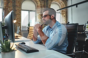 New ideas. Side view of young bearded man in eyeglasses and formal wear looking at computer and thinking while sitting