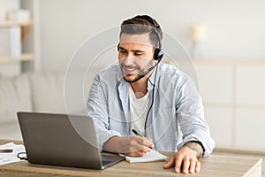 New knowledge online at home. Young man in headphones making notes and looking at laptop computer in living room