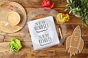 New mindset and new results text on notebook on wooden table with coffee cup and paper rocket. Business concept of positive