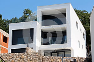 New modern apartment building with white facade and glass balcony fence with black window blinds surrounded with stone wall