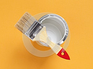 New natural bristle brush with wooden handle on the edge of an open white paint can over bright yellow textured background.