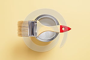 New natural bristle brush with wooden handle on an open white paint can over pastel yellow background. Construction painting work