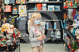 A new normal. Caucasian blonde girl in sanitary face mask shopping at toy store. Child wearing protective mask against coronavirus