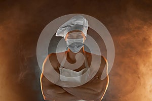 New normal concept. Muscular chef wearing protective medical mask, posing with folded arms on smoky background