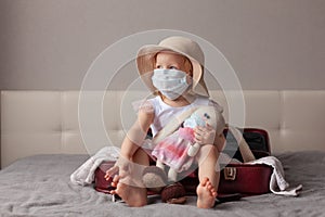 new normality travel, happy little girl sun hat and medical mask sits burgundy open suitcase, hugging toy knitted rabbit