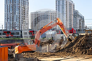 A new orange excavator is working on a construction site with a pile of brown soil. High-rise residential buildings and