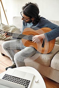 New skill. Young focused man sitting on sofa at home and playing acoustic guitar, watching online music course on laptop