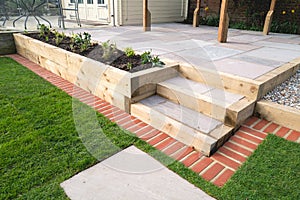 New steps in a garden or back yard leading to a raied patio, alongside a new raised flowerbed made using wooden sleepers. A