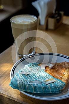 New trendy flat croissant with blue glaze lies on white plate in a cafe, next to coffee in a glass cup, vertical photo photo