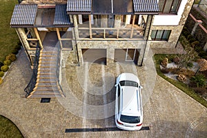 New white car parked in paved driveway with green lawn, decorative bushes and brick fence of big two story house with wide